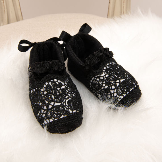 A pair of delicate black floral lace June Booties with white satin ribbon ties, placed on a soft white furry surface.