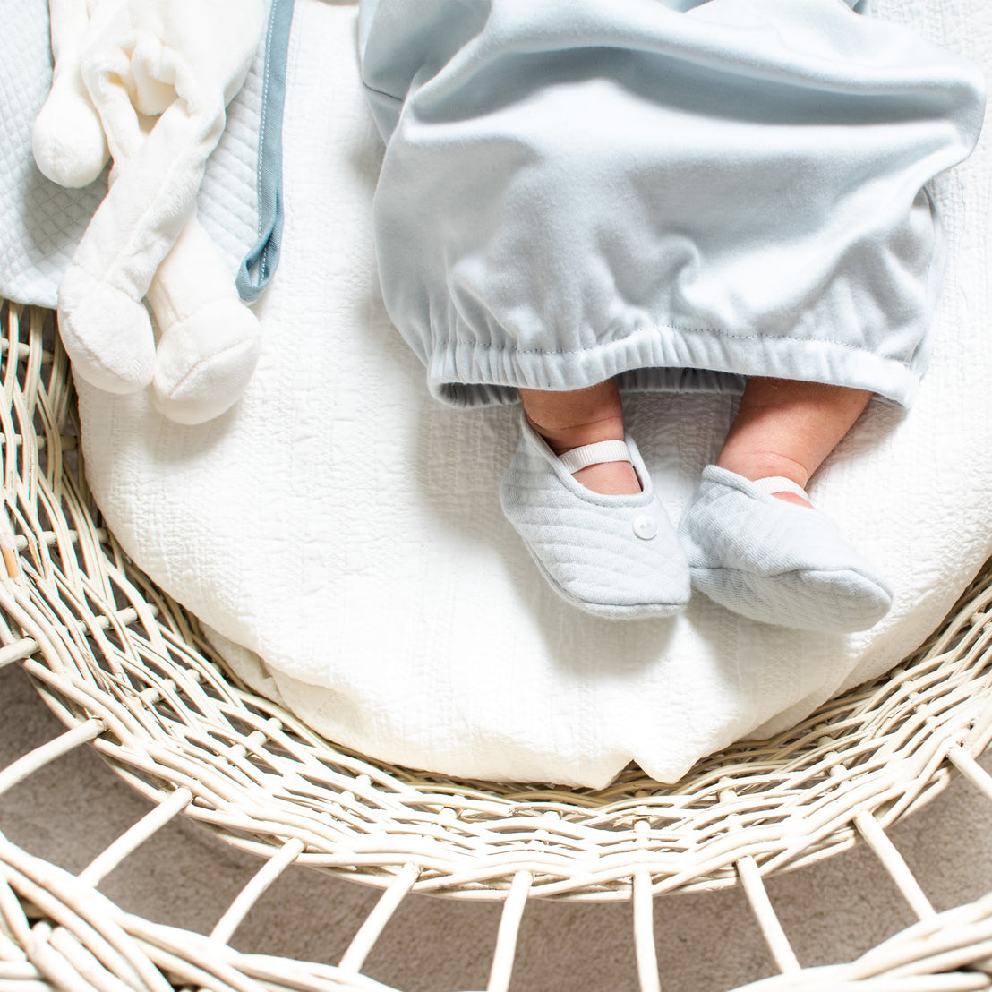 A newborn baby in a soft blue blanket with Aiden Quilted Booties lies asleep in a cozy, woven bassinet, demonstrating an upscale and serene nursery setting.