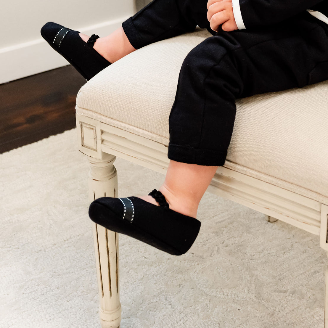 Baby boy sitting on a chair and wearing the James Booties. They are made from 100% Black French Terry Cotton featuring a stitched ribbon toe and a soft elastic strap.