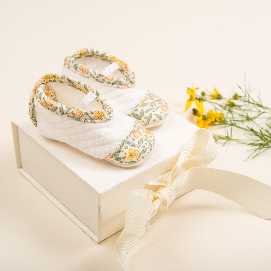 A pair of handcrafted, Petite Fleur Quilted Booties on a white box tied with a cream ribbon, placed beside yellow flowers on a light beige background.