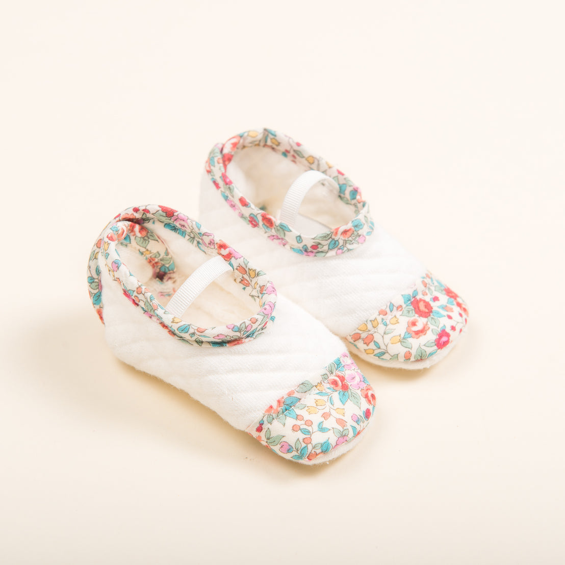 A pair of Petite Fleur Quilted Booties on a soft beige background. The shoes feature small floral prints in various colors and white straps across the tops.