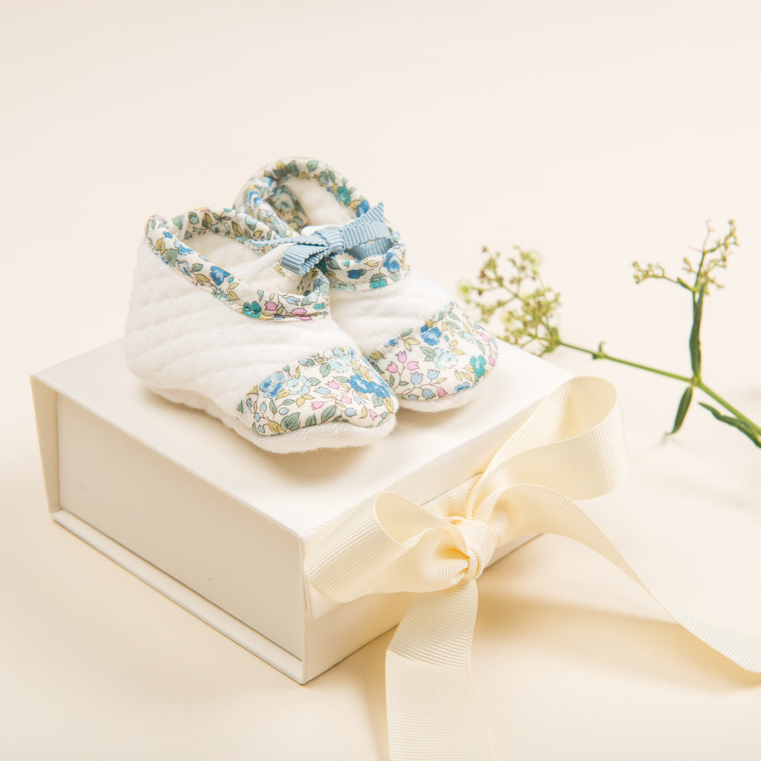 A pair of Petite Fleur Quilted Booties for newborn babies with floral pattern and blue ribbons displayed on a boutique white box adorned with a satin bow, accompanied by small white flowers on a light background.