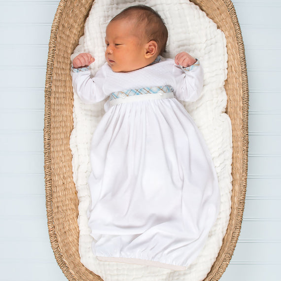 A newborn baby sleeps peacefully in a woven bassinet wearing a long white Mason Newborn Gown with light blue embroidery for a baptism, on a soft white blanket against a blue striped background.