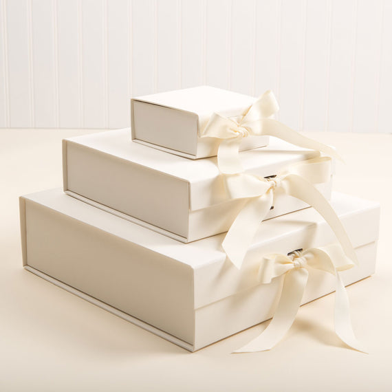 Three stacked Baby Beau & Belle gift boxes with magnetic closure and ivory satin ribbons on a pale background, ranging in size from small to large.