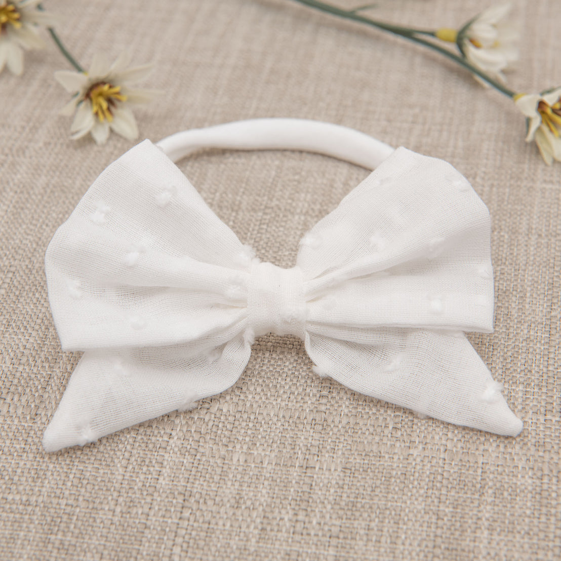 A Mila Bow Headband, a white Dotted Swiss cotton bow attached to a nylon headband, placed on a beige background and surrounded by small white daisies.