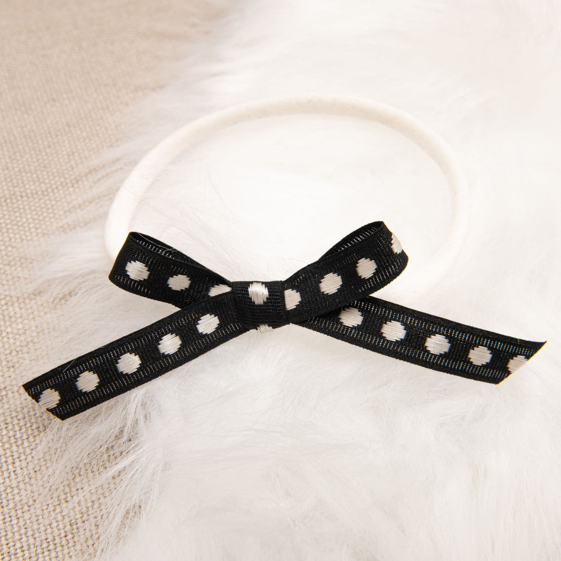 A black embroidered June Bow Headband with white polka dots tied into a bow on a white furry surface.