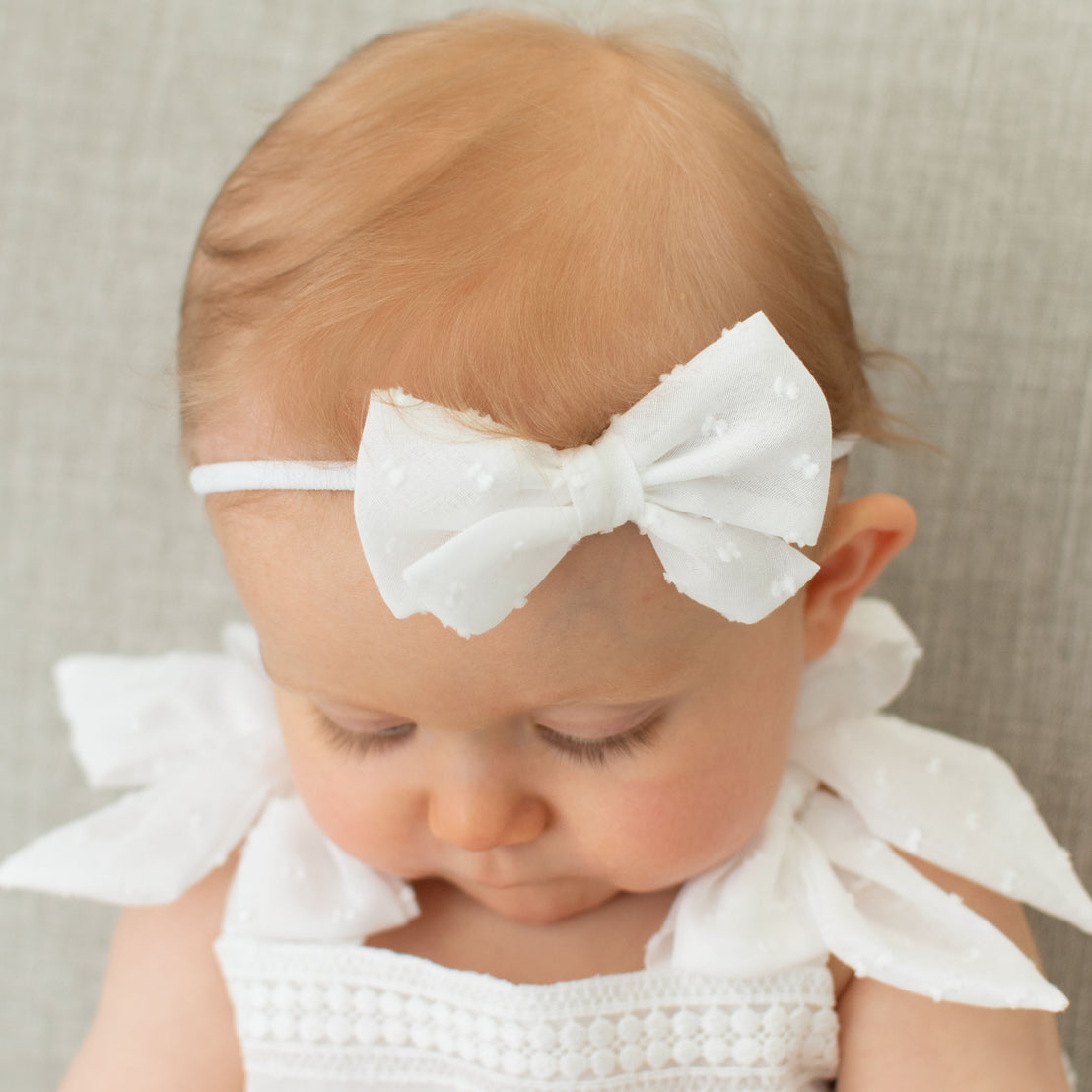 A newborn with light brown hair wearing the Mila Bow Headband and Mila Cotton Gown, looking downward against a textured grey background.