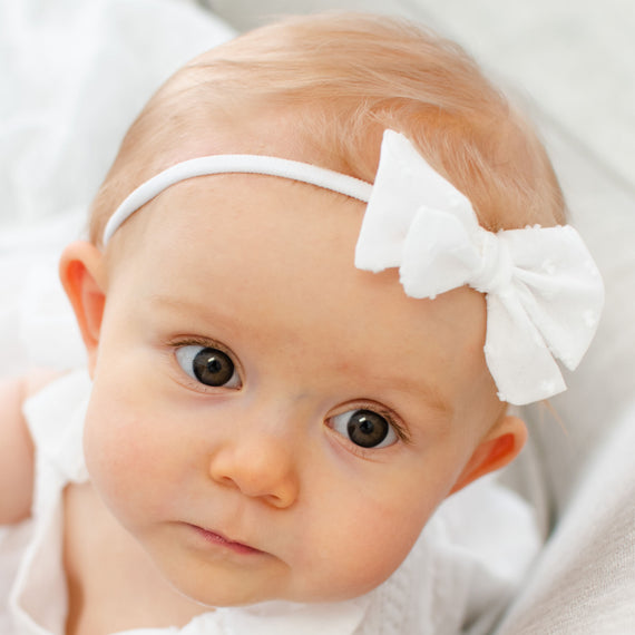 Close-up portrait of a baby with big brown eyes and light hair, wearing the Mila Bow Headband, a white Dotted Swiss cotton bow attached to a nylon headband, looking off to the side of the camera.