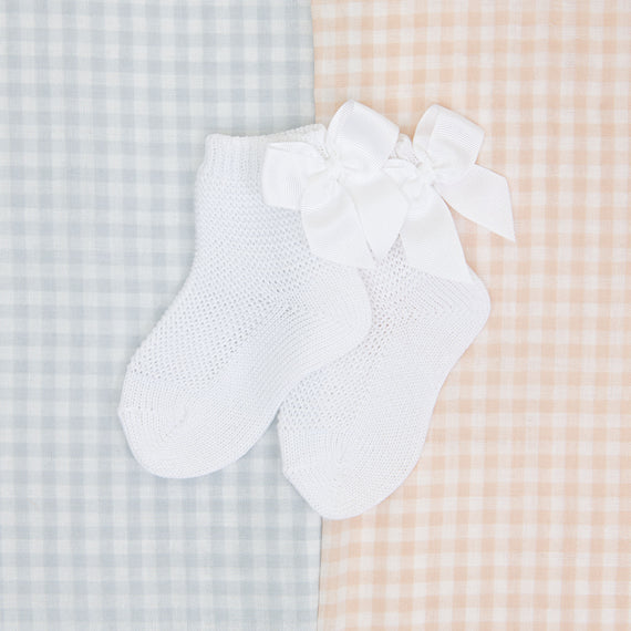 A pair of white newborn Garter Stitch Socks with Bow, displayed on a boutique gingham-patterned fabric background in soft blue and orange tones.