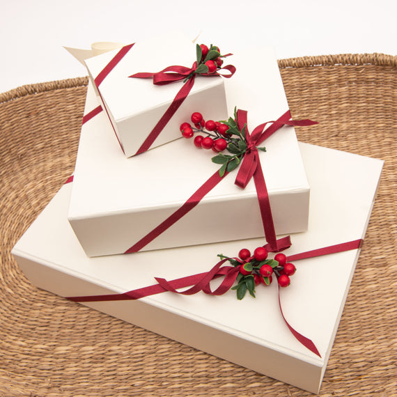 Three Ivory Gift Boxes with magnetic closures, red ribbons, and berry decorations are arranged in a wicker basket. 
Brand Name: Baby Beau & Belle