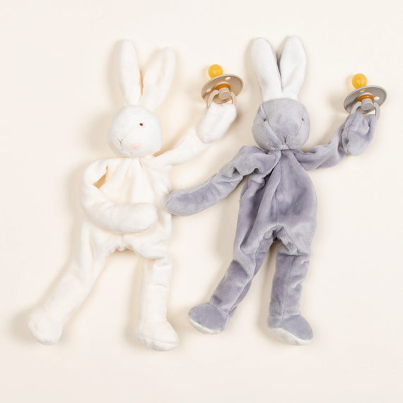 Two Silly Bunny Buddy & Pacifier toys, one white and one gray, designed for newborns, each holding a pacifier against a light background. These bunnies appear soft and are ideal for boutique settings.