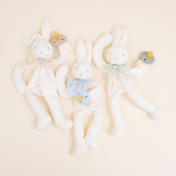 Flat lay photo of three Theodore Bunny Buddy Pacifier Holders. Stuffed animal floppy bunny made from a soft velour. Available colors include blue, green, and tan/white.