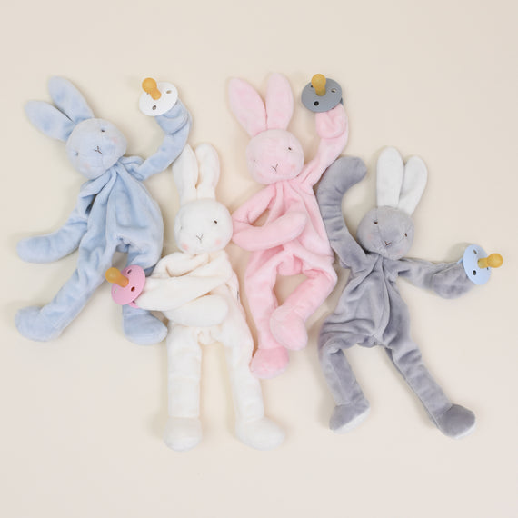 Five Silly Bunny Buddy | Pacifier Holder toys in blue, grey, white, pink, and gray, each holding a small pacifier and arranged symmetrically on a light beige background, exuding an upscale vintage charm.