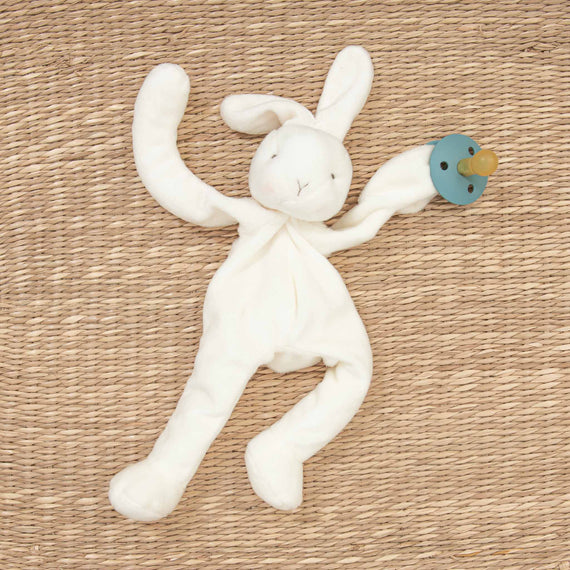 Aiden Silly Bunny Buddy & Pacifier toy with a pacifier attached lies on a woven mat. The toy has floppy ears and an upscale, serene expression, creating a calm and comforting scene.