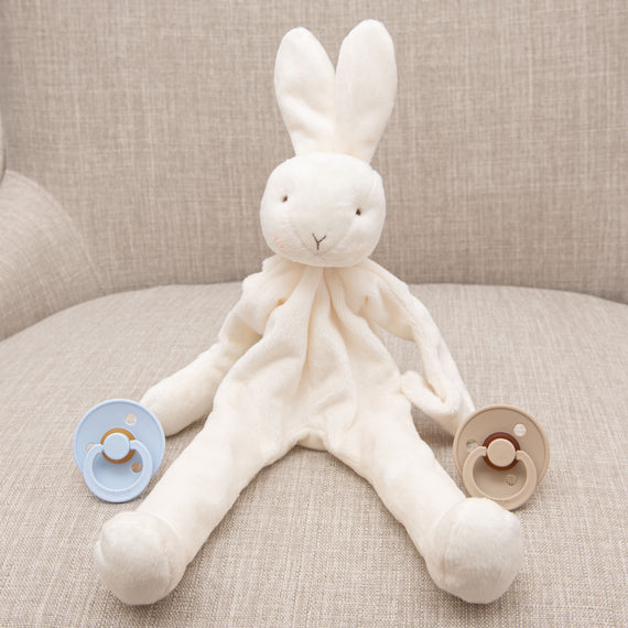 A Isla Silly Bunny Buddy & Pacifier sits on a beige sofa, flanked by two pacifiers, one blue and one beige, placed on either side of it.