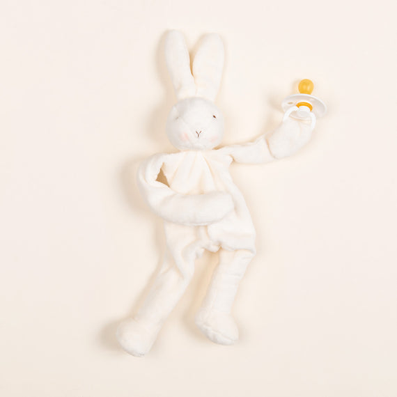 A plush bunny doll holding a bibs pacifier in ivory color. Set against a beige backdrop. 