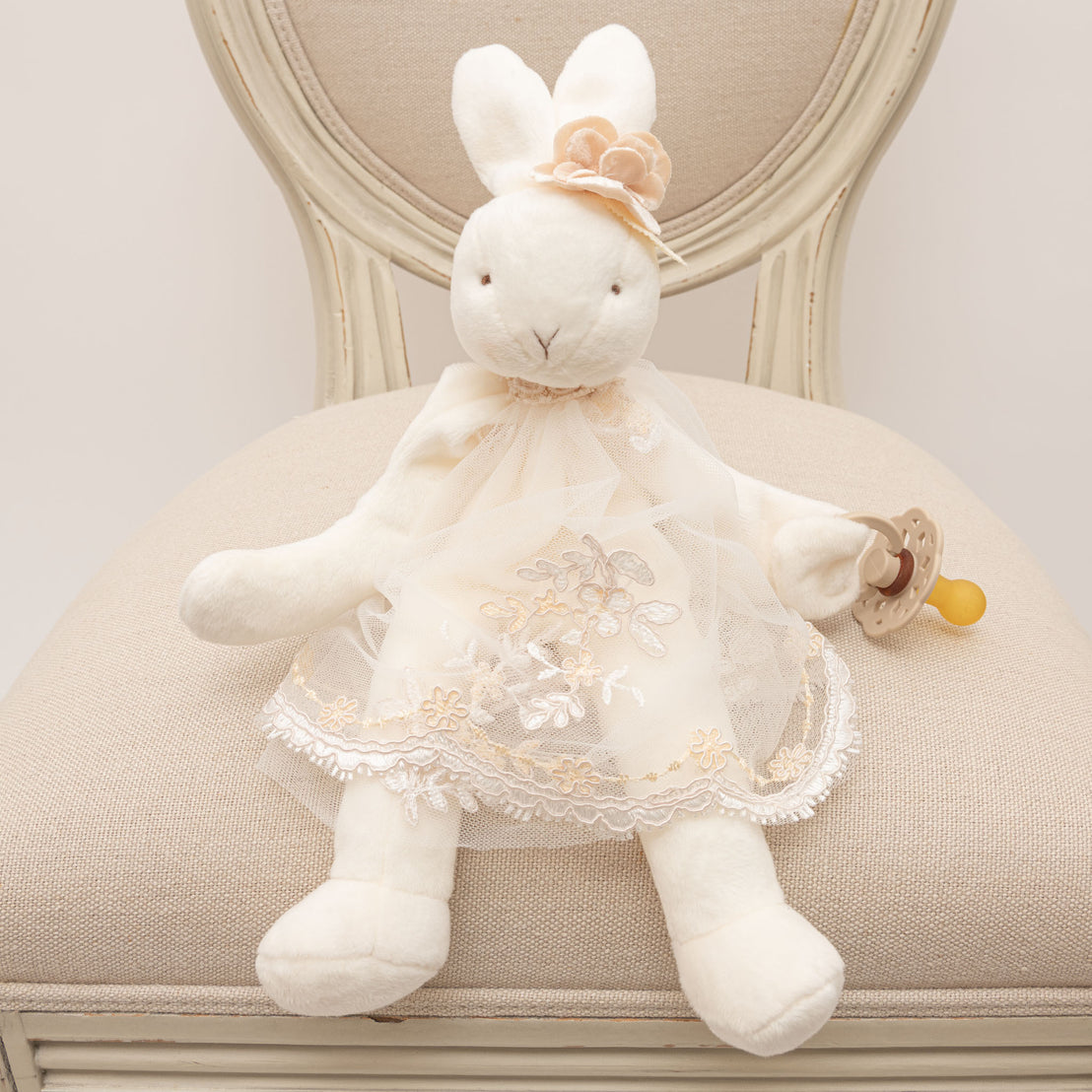 A Kristina Silly Bunny Buddy dressed in a delicate white dress with floral embroidery, sitting on a beige chair, holding a small pacifier, perfect for a christening.