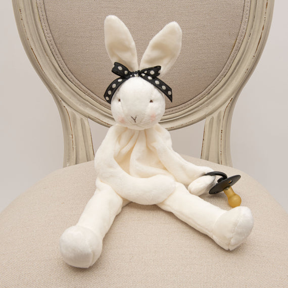 A June Silly Bunny Buddy pacifier holder with a black polka-dot bow sits on a chair, holding a baby's pacifier. The bunny, crafted from soft velour, has long ears.