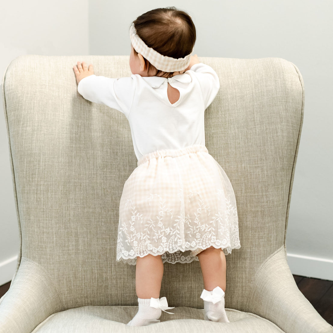 A toddler in an Isla Bubble Skirt Set and matching Isla Tie Headband stands on a beige armchair, facing away from the camera and holding onto the chair's back.