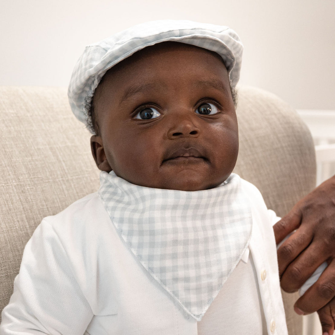 A baby dressed in a traditional white outfit with an Isla Bandana Bib and a vintage light hat looks curiously at the camera.