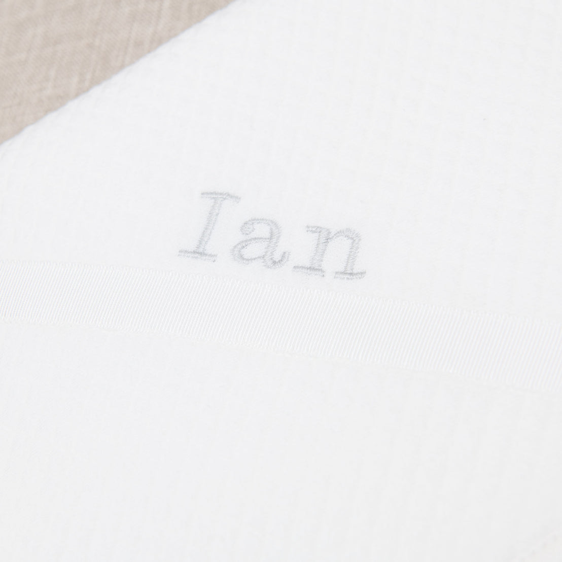 Close-up of a white fabric with the name "Ian" embroidered in gray thread, centered above a horizontal satin ribbon on the Ian Personalized Blanket in a traditional designer style.