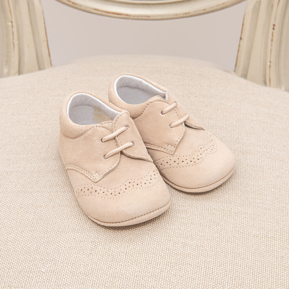A pair of small camel suede shoes for toddlers placed neatly on a beige cushioned chair with a classic wooden frame in the background.