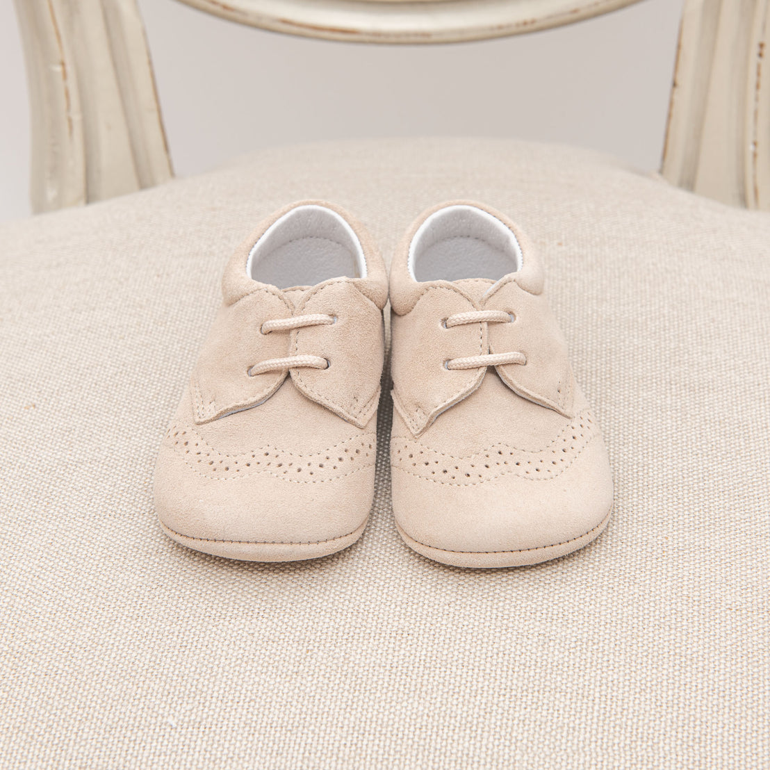A pair of small beige handmade Camel Suede Shoes with laces, placed neatly on a textured cream cushion and set against a vintage-style wooden chair.