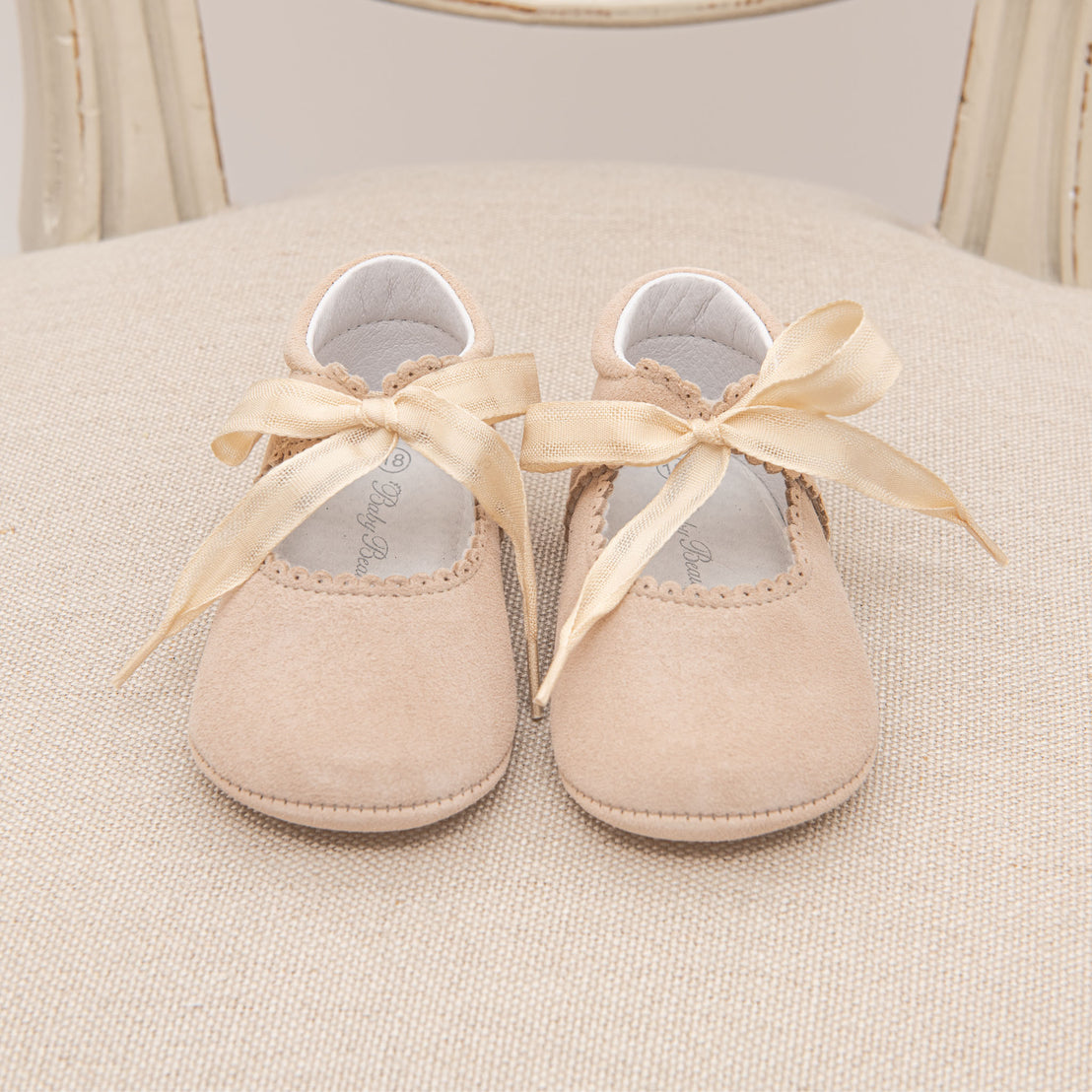 A pair of small beige Kristina Tie Mary Janes with yellow ribbons on a beige fabric chair, presenting a charming and vintage-inspired appearance.