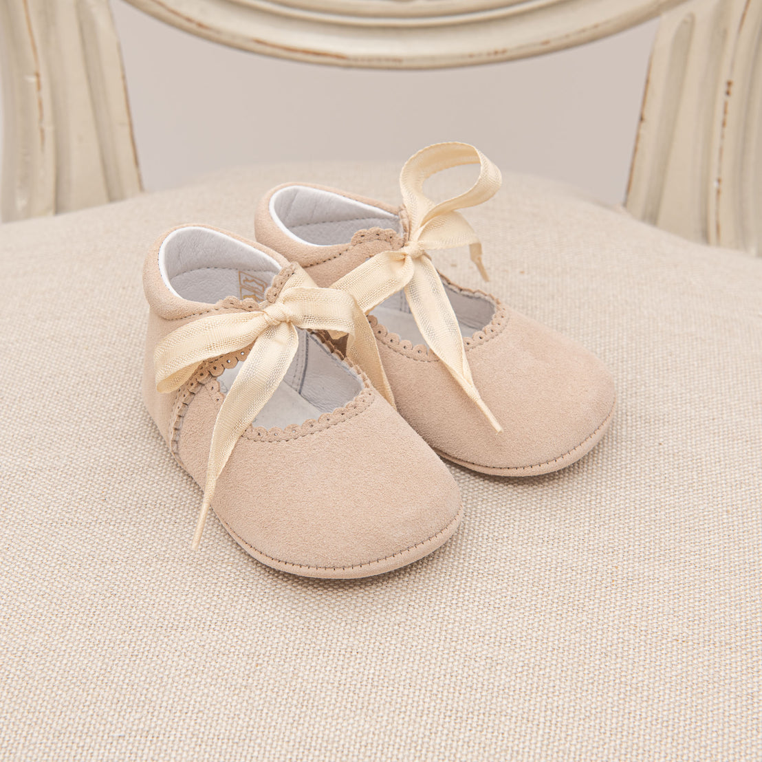 Pair of Kristina Tie Mary Janes with ribbon ties on a white cushioned chair, showcasing soft, suede-like texture and delicate lace details for a christening.
