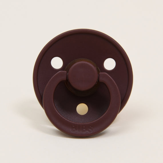 A Chestnut-colored pacifier with a round shield and two large air holes, featuring a handle and a natural rubber nipple, against a plain light background.