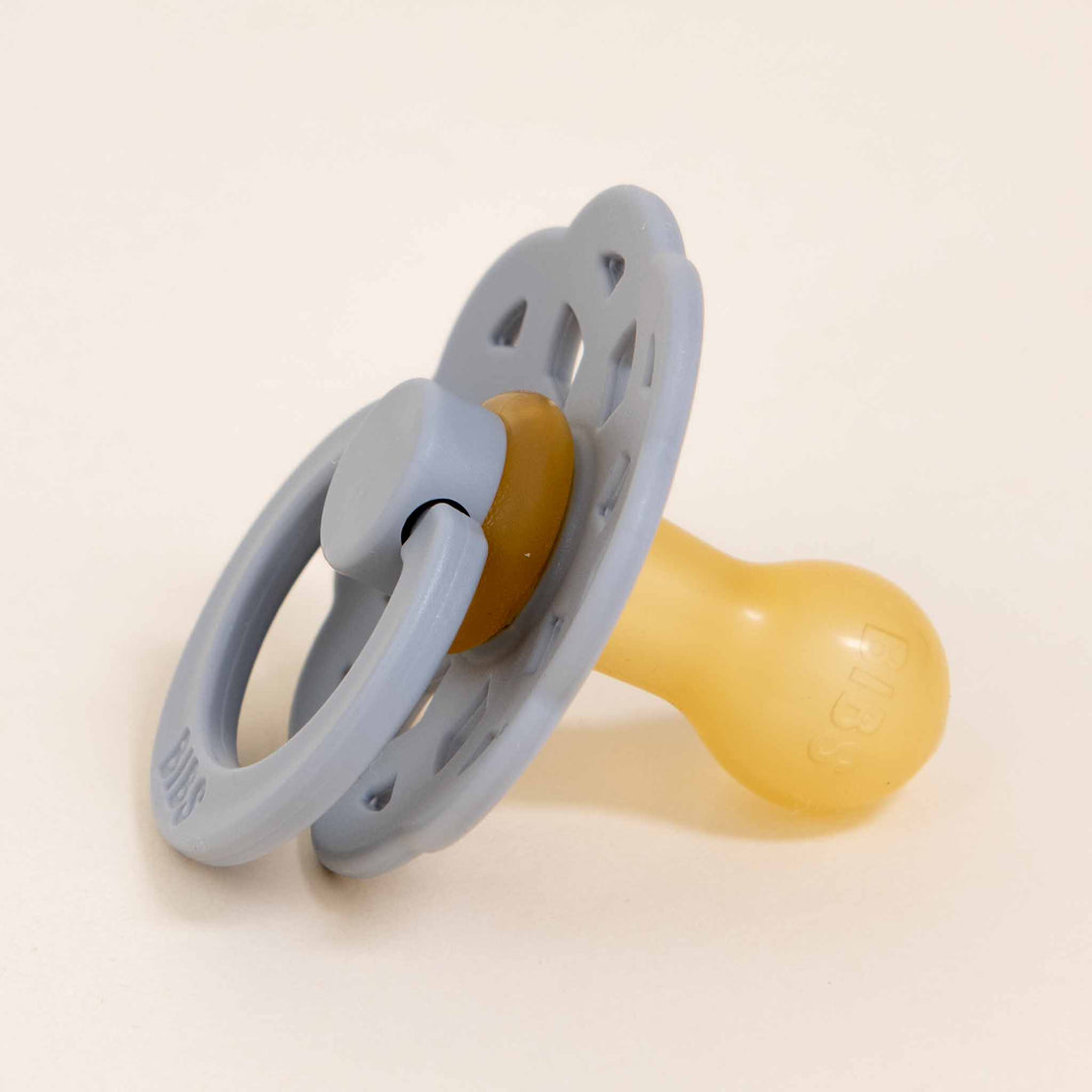 A Bibs Lace Pacifier in Cloud with a gray handle and yellow nipple, placed on a light beige background.