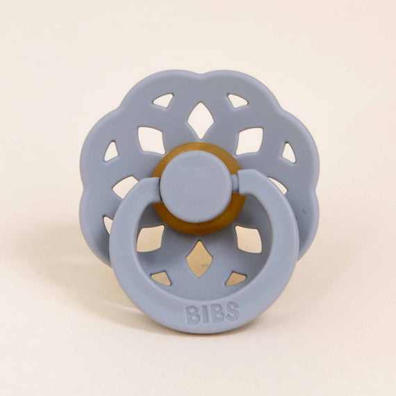 A pale blue, flower-shaped silicone pacifier with a wooden button center labeled "Bibs Lace Pacifier 2 Pack | Cloud" on a beige background, designed and manufactured in Denmark.