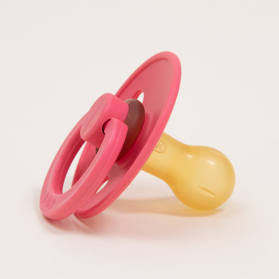 A close-up image of a pink Bibs Pacifier in Coral with a translucent yellow nipple and a circular handle on a white background.