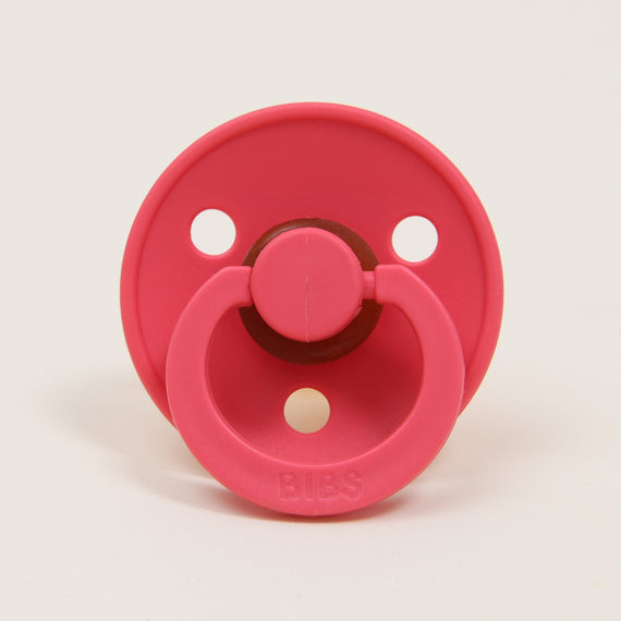 A Bibs Pacifier 2 Pack | Coral with a circular shield and two air holes, featuring a handle and a natural rubber nipple, with the brand name "bibs" embossed on the front.