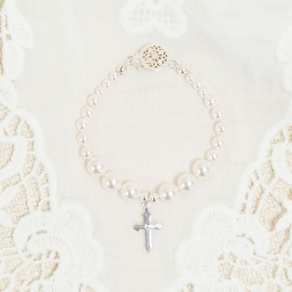 A flat lay photo baby jewelry: the white luster pearl bracelet with a silver cross in the middle. Photographed on top of the Adeline Christening Gown.