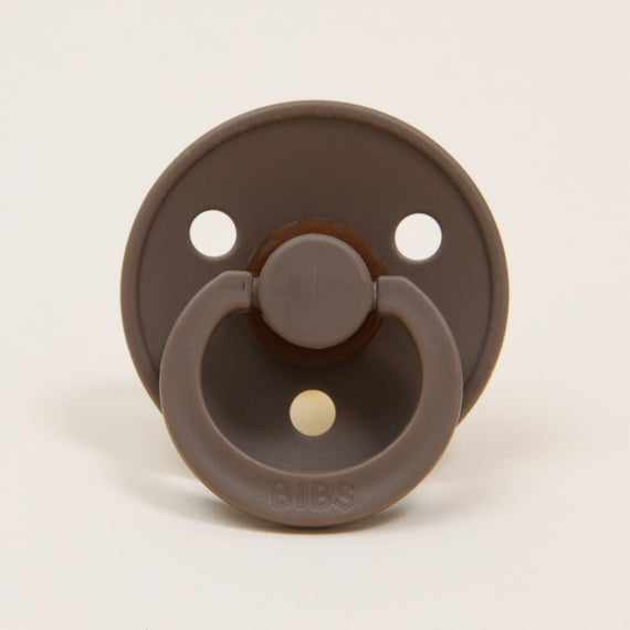 A Bibs Pacifier 2 Pack | Dark Oak featuring a round shield with three holes for ventilation and a handle, set against a plain white background.