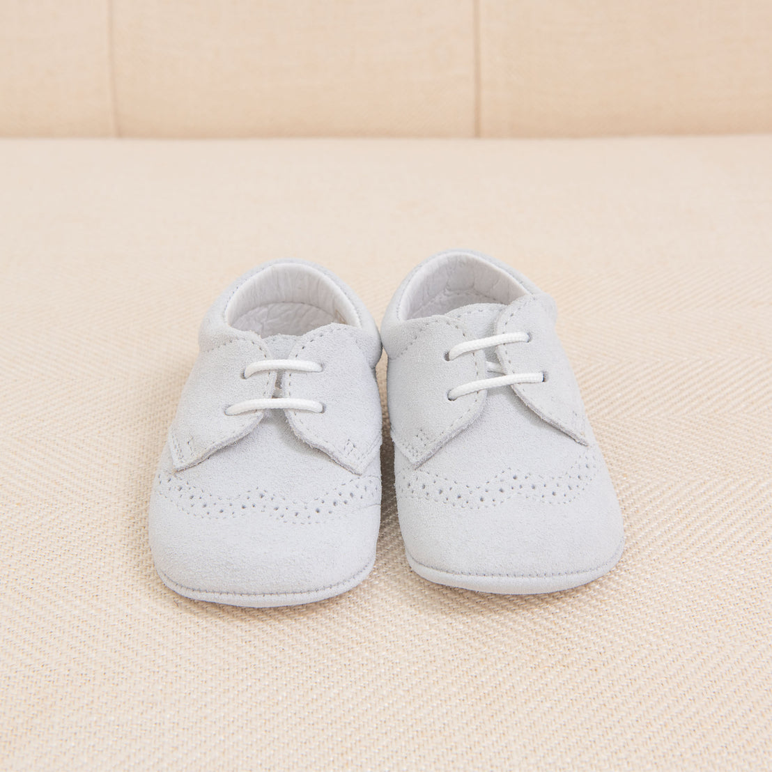A pair of light gray Baby Beau & Belle Boys Suede Shoes with laces, set on a beige fabric background.