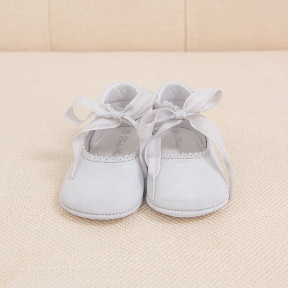 A pair of small Dove Grey Suede Tie Mary Janes with ribbon ties on a cream-colored fabric background.