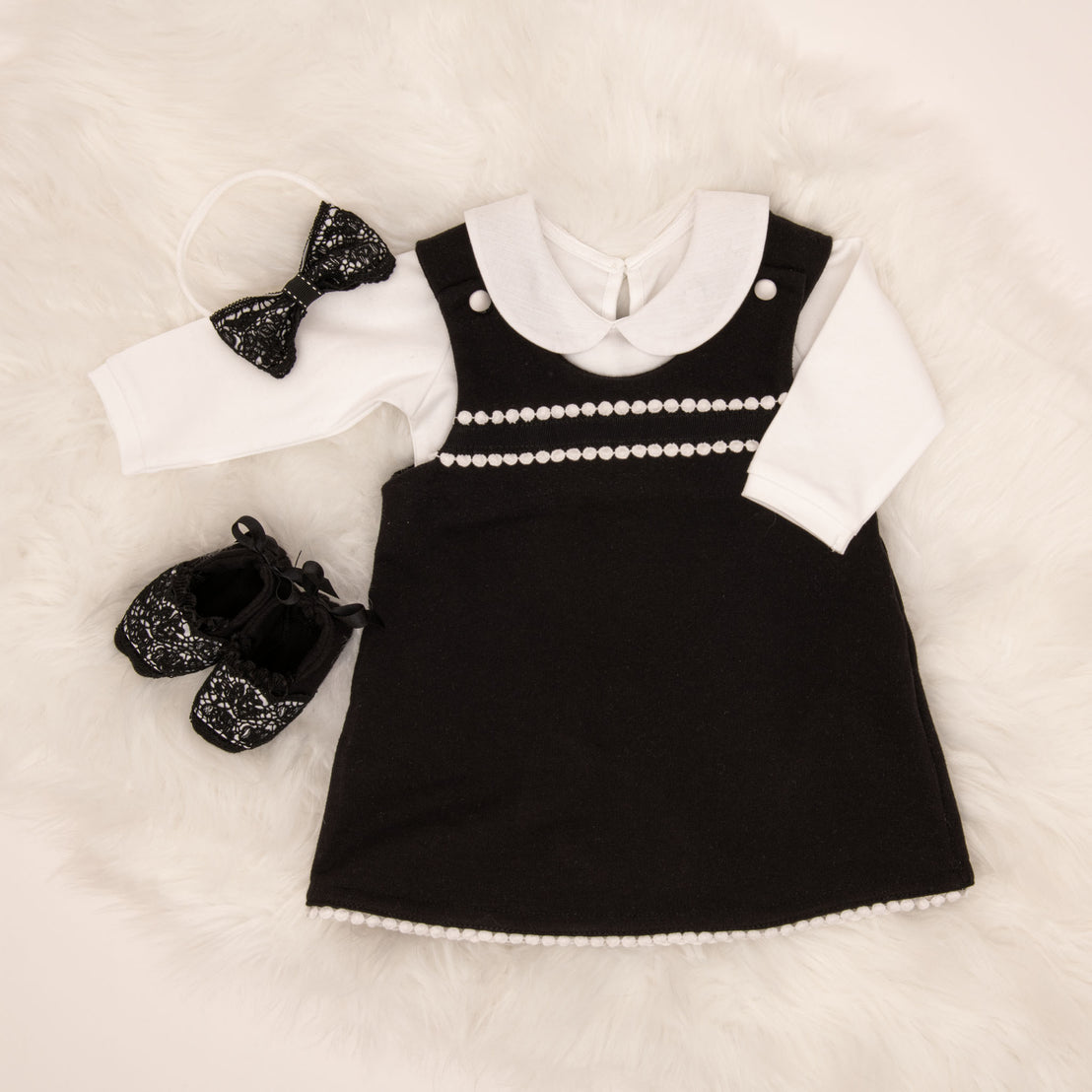 A stylish baby girl outfit on a fluffy white background, including a June Jumper Dress Set with white pearl details, matching black and white shoes, and a lace headband with bow.