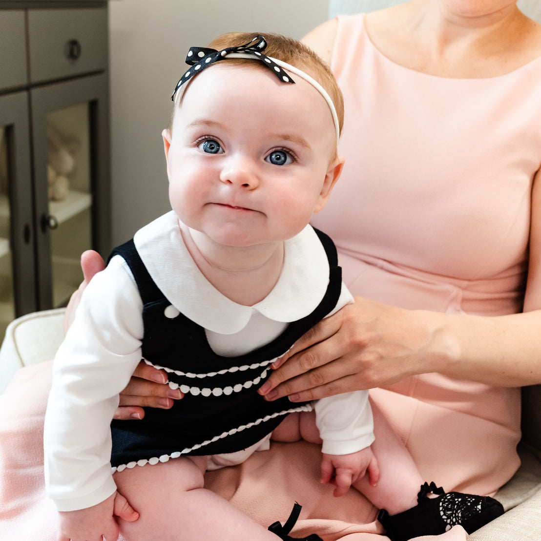 A baby with blue eyes and wearing a headband and black floral lace dress looks directly at the camera, held by a woman in a pink blouse, partially visible.