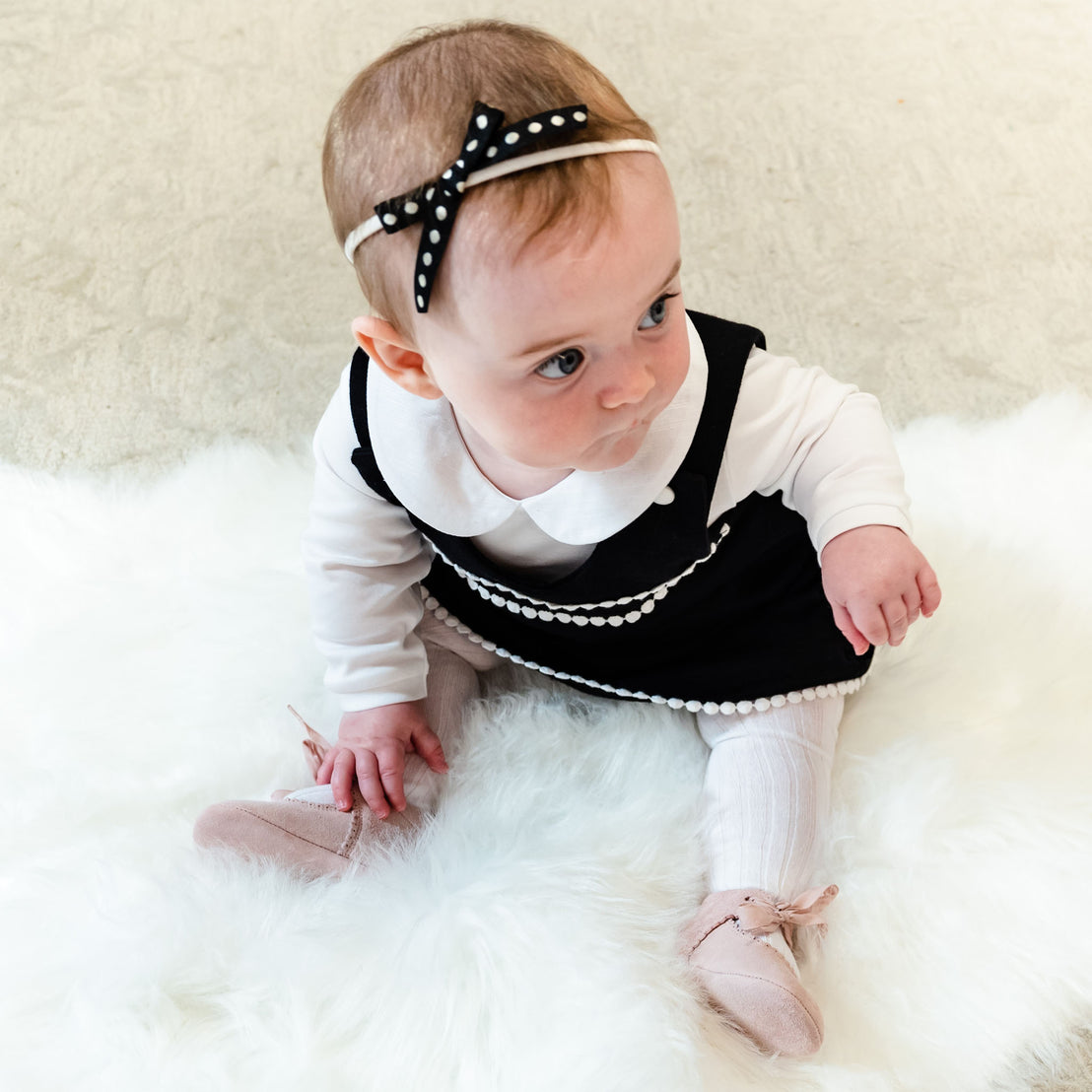 A baby girl in a stylish black and white dress with a June Bow Headband sits on a fluffy white rug, looking curiously to the side. She wears pink shoes and white socks.