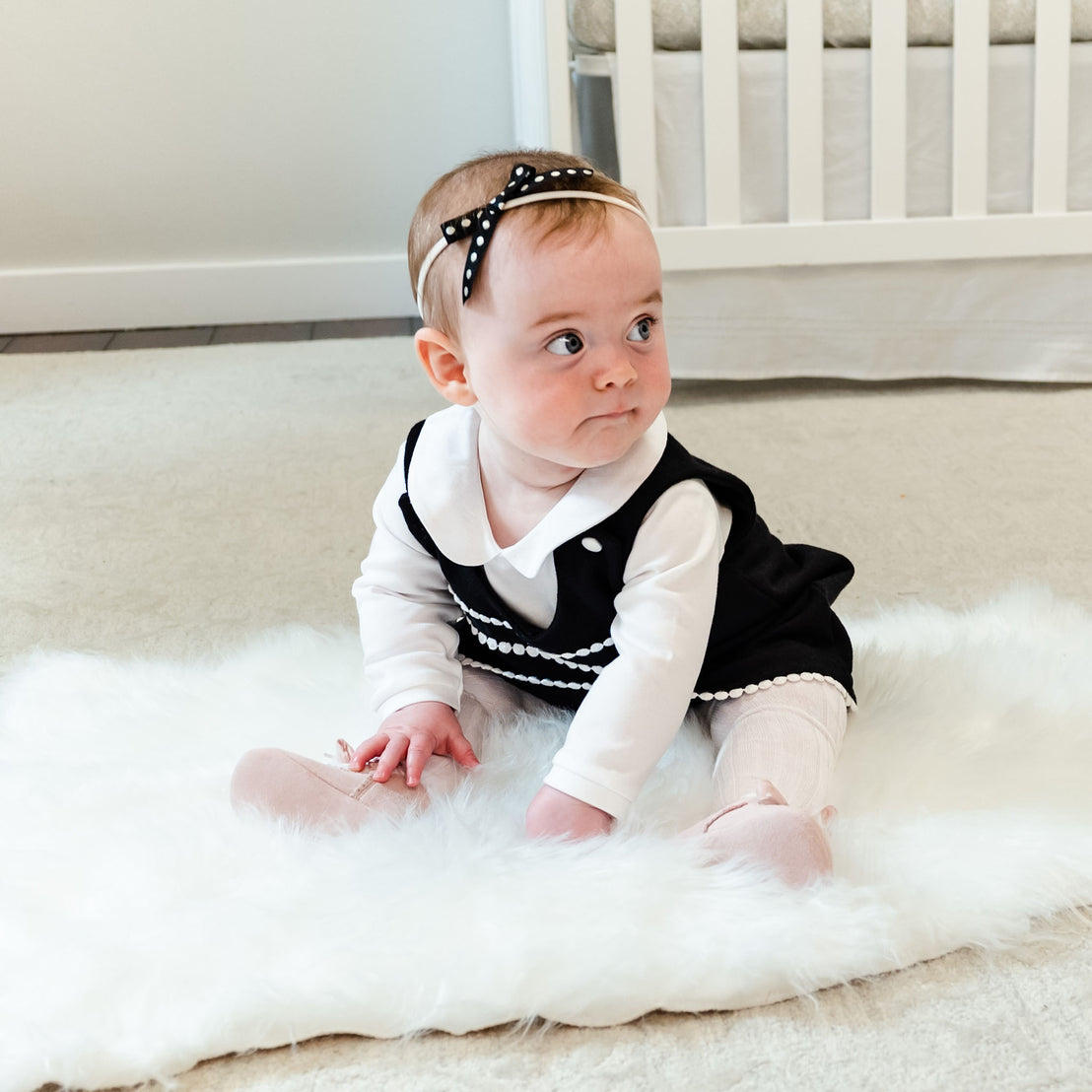 A baby girl sits on a plush rug, wearing a black and white dress with pink leggings and June Suede Tie Mary Janes, gazing thoughtfully to the side, with a crib partially visible.