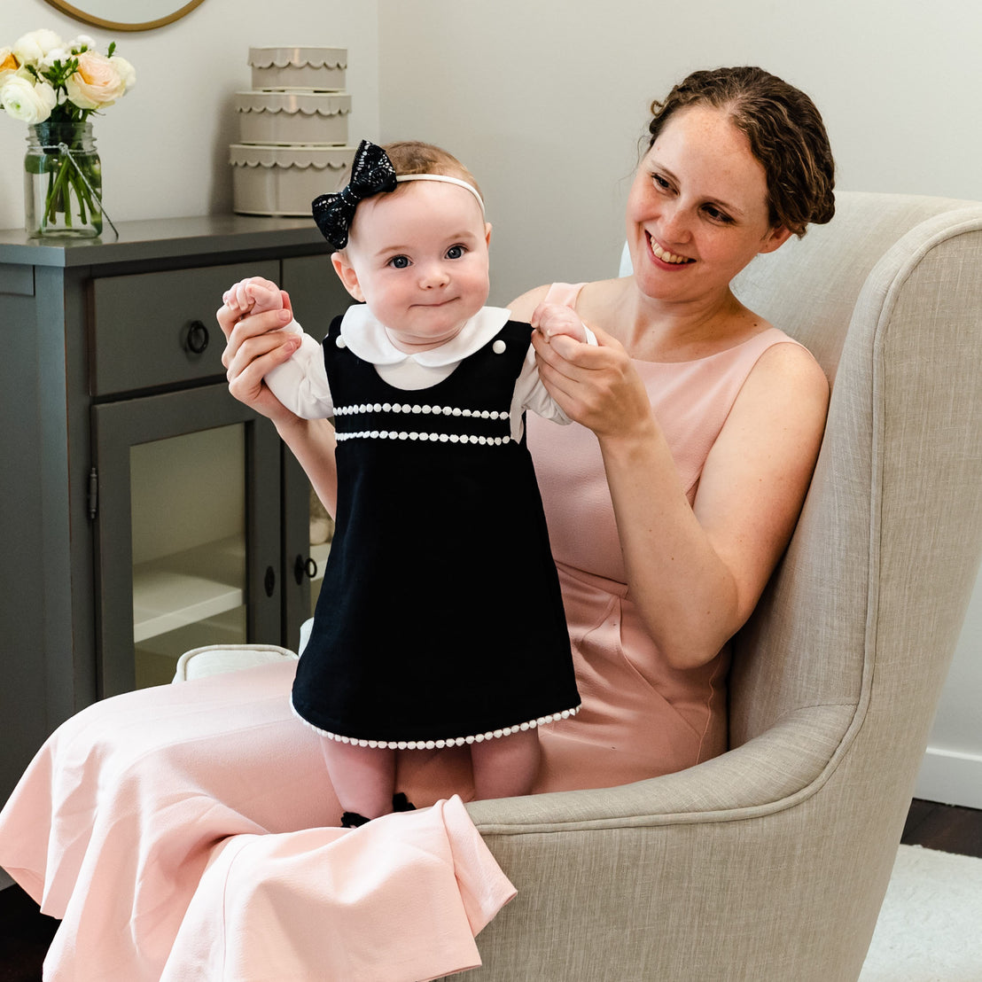 A woman in a pink dress smiles, sitting in an armchair and holding up a baby girl in a June Jumper Dress Set with pearl trim, who is playfully trying to stand. 