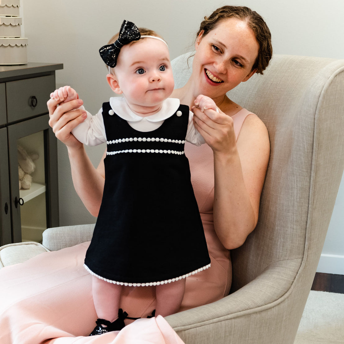 A smiling woman sits in a beige armchair, holding a baby girl dressed in a black floral lace outfit with a June Lace Bow Headband. 