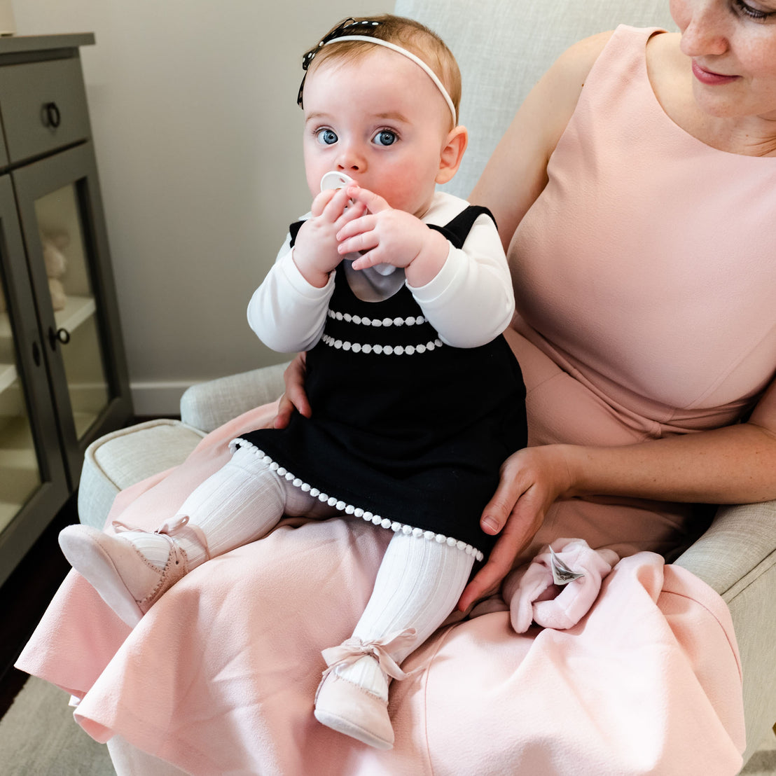 A mother in a pink dress holds her baby girl, wearing a black and white dress with a pearl trim and white socks, sitting on her lap. The baby is chewing on her pacifier and also wearing June Suede Tie Mary Janes.