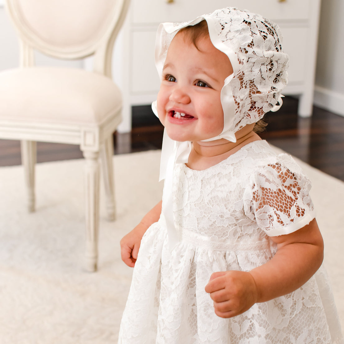 A joyful toddler in a traditional white lace dress and Rose Lace Bonnet smiles while standing on a hardwood floor with a vintage white chair in the background.