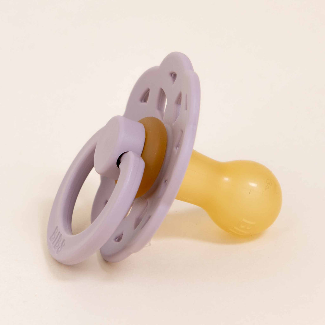 A Bibs Lace Pacifier in Dusky Lilac with a lavender shield and handle, featuring a yellow nipple, designed in Denmark, set against a light beige background.