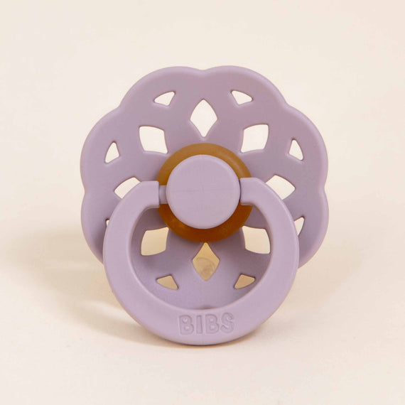 A Bibs Lace Pacifier in Dusky Lilac with a circular shield and symmetric ventilation holes, featuring a brown handle and button on a neutral background.