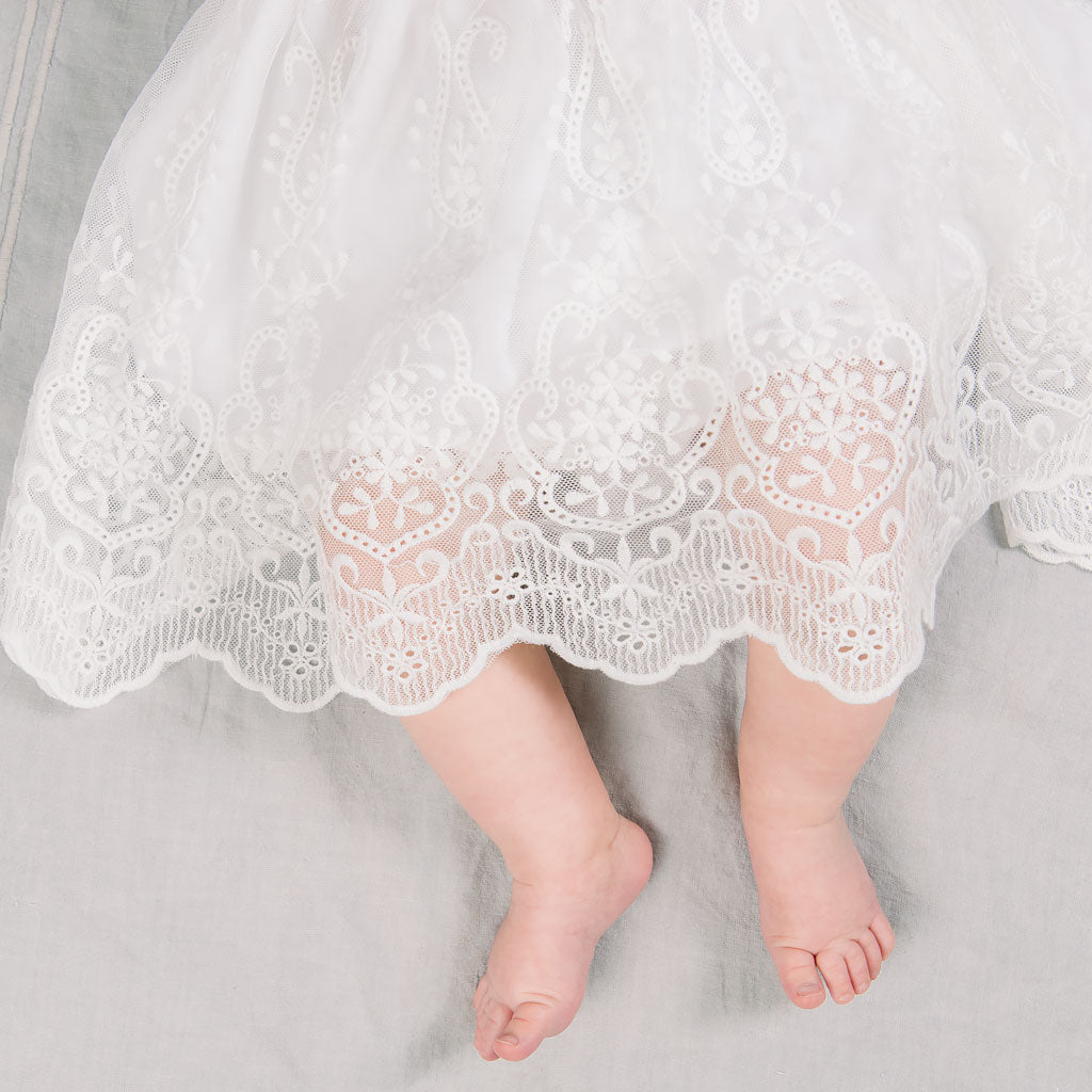 A close-up of a baby's bare feet peeking out from under an Eliza Lace Dress & Headband with vintage patterns, set against a soft grey background during an upscale christening event.