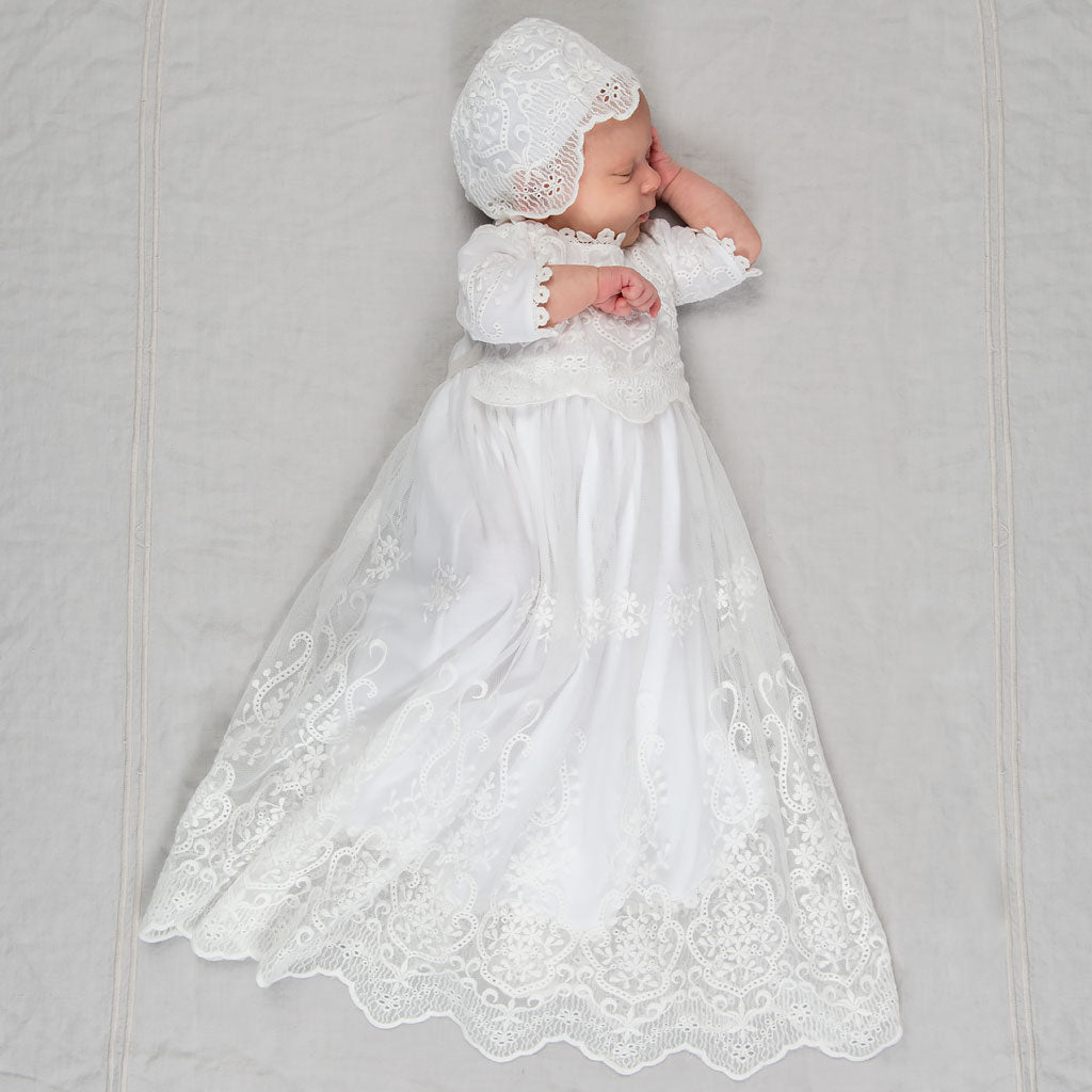 A newborn baby sleeps on a soft gray background, dressed in the Eliza Blessing Gown & Bonnet with intricate heirloom lace detailing.